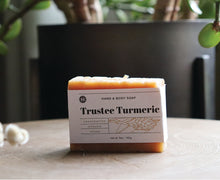Load image into Gallery viewer, Trustee Turmeric hand and body soap bar on a stand with plant in background. handcrafted, organic, vegan. Olive Seed